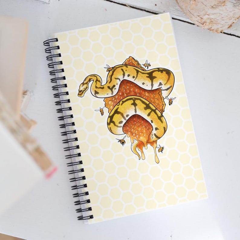A spiral-bound notebook with a pale yellow honeycomb-patterned cover, featuring an illustration of a yellow and black snake wrapped in orange honeycomb with bees, photographed on a white surface.