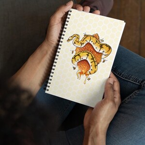 A person holding a spiral-bound notebook with a yellow and orange snake coiled around a piece of honeycomb with bees  on the cover, shown from above while seated wearing blue jeans.
