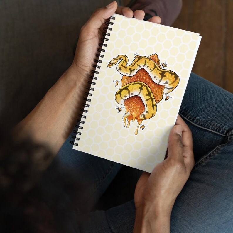 A person holding a spiral-bound notebook with a yellow and orange snake coiled around a piece of honeycomb with bees  on the cover, shown from above while seated wearing blue jeans.