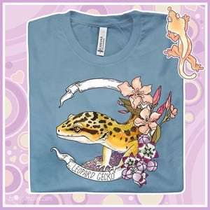 Könnte beinhalten: Ein hellblaues T-Shirt mit einem Aufdruck eines Leopardgeckos, umgeben von Blumen. Der Gecko ist gelb und schwarz mit weißem Hintergrund. Der Text "LEOPARD GECKO" ist auf einem weißen Banner geschrieben.
