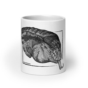 Pode incluir: Uma caneca de cerâmica branca com uma ilustração detalhada em preto e branco de uma cabeça de lagarto com a língua de fora. A arte é emoldurada por uma borda preta fina e inclui o texto científico Salvator Merianae na parte inferior.