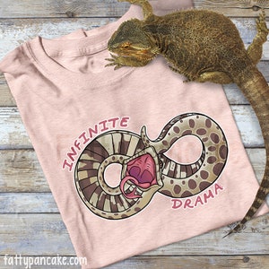 May include: Pink t-shirt with a brown and white snake design in the shape of an infinity symbol. The snake has its mouth open and is biting its tail. The text "INFINITE DRAMA" is printed on the shirt.