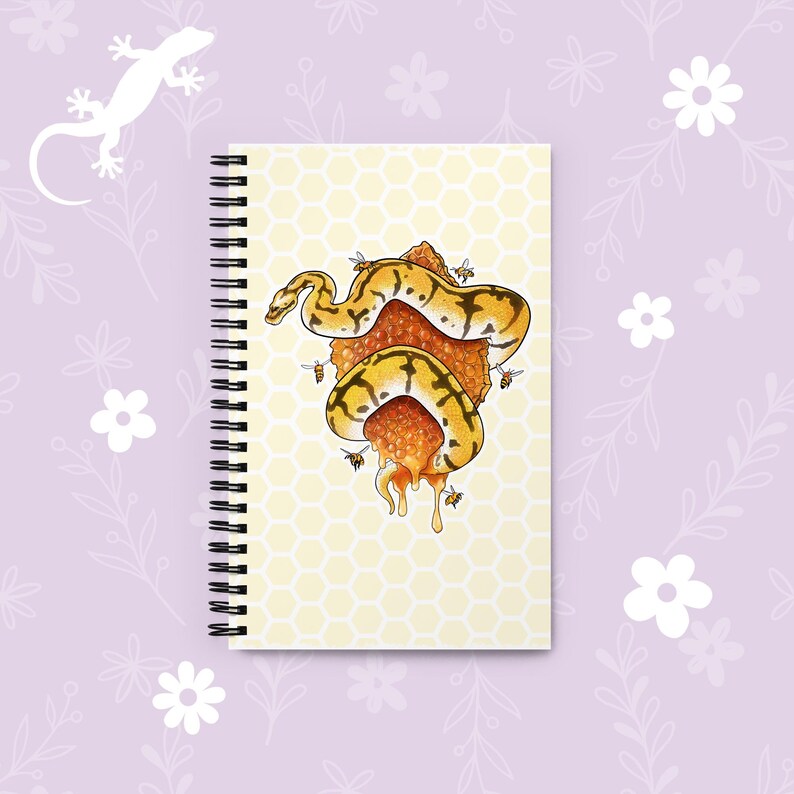 A spiral-bound notebook with a pale yellow honeycomb pattern cover, featuring an illustration of a yellow and black snake wrapped around a honeycomb structure. The notebook is displayed on a light lavender background decorated with white floral graphics and a white lizard silhouette.