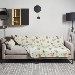 May include: A light green fleece throw blanket with a repeating pattern of colourful doughnuts with sprinkles and green leaves. The blanket is folded and draped over a light grey sofa.