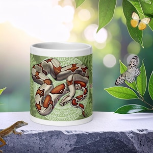 Könnte beinhalten: Ein weißer Keramikbecher mit einer detaillierten Illustration einer zusammengerollten Boa Constrictor mit braunen und cremefarbenen Zeichnungen vor einem hellgrünen kreisförmigen Mandalamuster und Blattmotiven, mit dem roten Textfragment Boa.