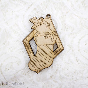 Pode incluir: Um enfeite de Natal de madeira com um gecko de desenho animado espiando de uma meia listrada. O enfeite tem a forma de um diamante e tem um furo na parte superior para pendurar.