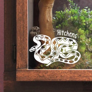 Könnte beinhalten: Ein weißer Vinyl-Aufkleber mit einer stilisierten Schlangenillustration und dem Text Hitchens auf einer Glasoberfläche. Das Design zeigt eine gewundene Schlange mit komplexen Mustern auf ihrem Körper, platziert über einem Holzrahmen und grünem Laub.