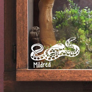 May include: A white vinyl decal applied to a glass surface featuring a stylised snake with a spotted pattern and the name Mildred written in a rounded sans-serif font below the snake. The decal is positioned on a wooden enclosure frame.