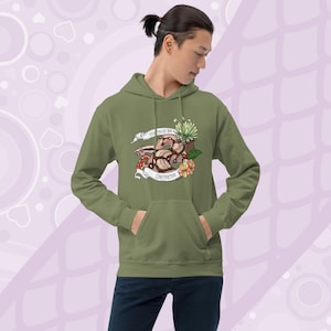 Puede incluir: Sudadera con capucha verde oliva con bolsillo delantero. La sudadera presenta un gráfico de una serpiente, flores y el texto "Red Tailed Boa" y "Constrictor" en una pancarta. La persona lleva vaqueros azul oscuro.