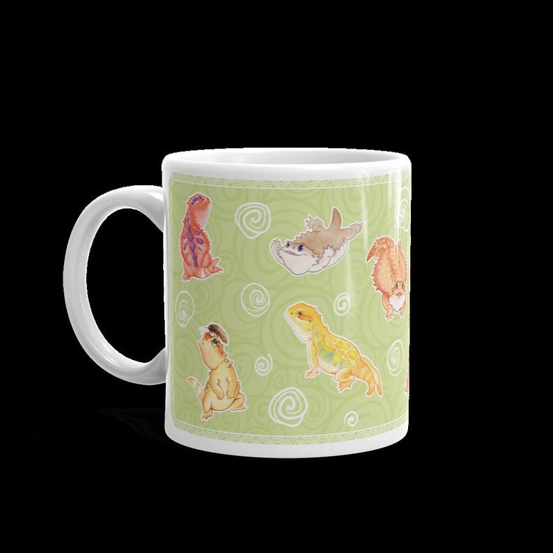 May include: White ceramic mug with a green background and a repeating pattern of cartoon bearded dragons and a fish. The bearded dragons are in various poses and colors, including yellow, orange, and red.