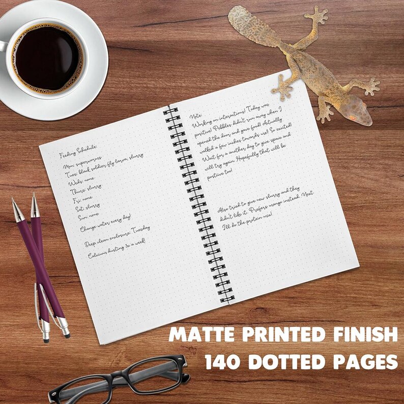 An open spiral-bound notebook with a matte white page finish lies on a wooden desk, surrounded by a cup of black coffee, two purple pens, eyeglasses, and a leaf tailed gecko. Large text at the bottom reads 'MATTE PRINTED FINISH, 140 DOTTED PAGES'.