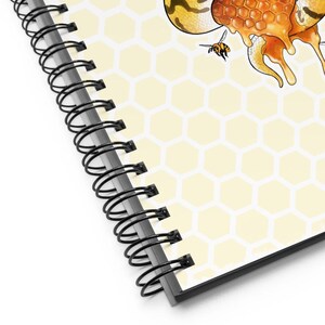 A close-up of a spiral-bound notebook showing the cover design with a pale yellow honeycomb pattern, dripping orange honey, a bee, and part of a floral illustration. The notebook has a black spiral binding and is photographed on a white background.