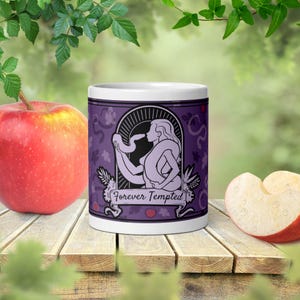 Könnte beinhalten: Ein weißer Keramikbecher mit einem violetten Grafikdesign, das eine Figur mit einer um den Körper gewickelten Schlange zeigt. Die Illustration ist von einem Torbogen umrahmt und enthält den Text Forever Tempted auf einem Banner mit Äpfeln.