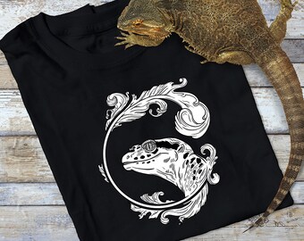 Może przedstawiać: Czarny T-shirt z białym nadrukiem przedstawiającym jaszczurkę w okularach przeciwsłonecznych w ozdobnym kółku. Jaszczurka jest zwrócona w prawo i ma szczegółowy wzór. Koszulka jest złożona, a na niej leży jaszczurka.