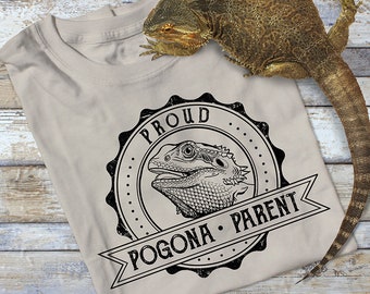 Op de afbeelding: Een beige T-shirt met een zwart-witte afbeelding van een baardagame. De afbeelding is een ronde stempel met de tekst "Proud Pogona Parent".
