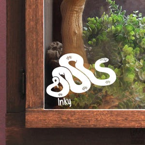 May include: A white vinyl decal featuring a stylised snake illustration with the text Inky written in a white sans-serif font below the snake. The decal is applied to the glass surface of a wooden enclosure containing green foliage.