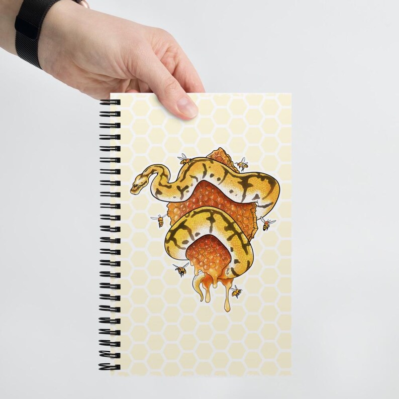 A hand holds a spiral-bound notebook against a light gray background. The notebook cover has a pale yellow honeycomb pattern and features an illustration of a yellow and black snake wrapped around a honeycomb with dripping honey.