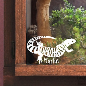 May include: A white vinyl decal of a lizard with a striped tail and body, featuring the name Marlin in a sans-serif font, applied to the glass front of a wooden enclosure containing a textured branch and green foliage.