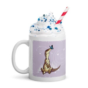 May include: A white ceramic mug featuring a graphic of a brown spotted lizard sitting on the ground with a small blue butterfly on its nose. The mug is filled with white whipped cream topped with blue sprinkles and a red and white striped straw.