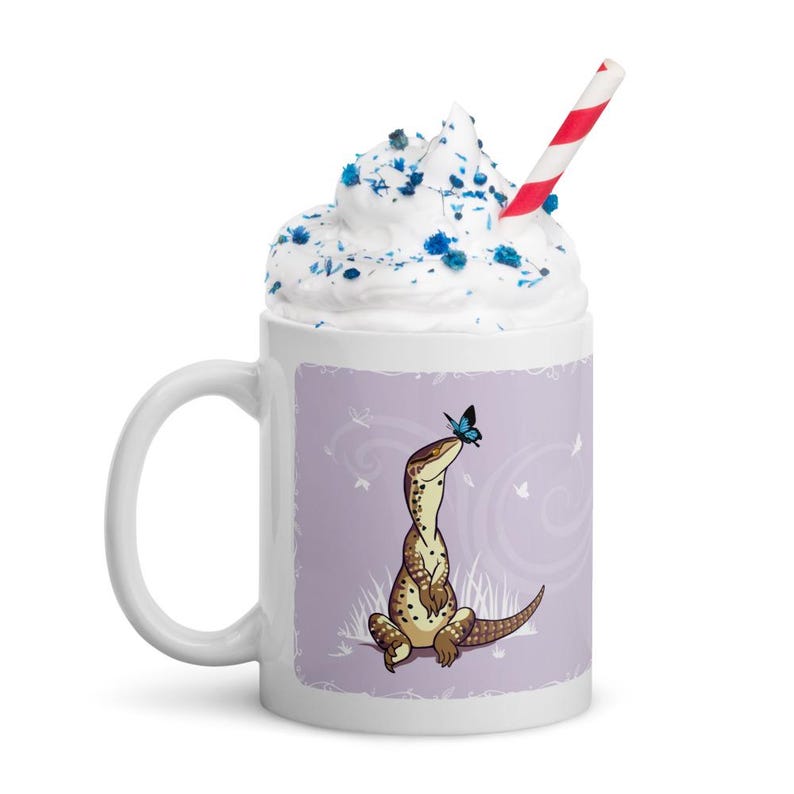 May include: A white ceramic mug featuring a graphic of a brown spotted lizard sitting on the ground with a small blue butterfly on its nose. The mug is filled with white whipped cream topped with blue sprinkles and a red and white striped straw.
