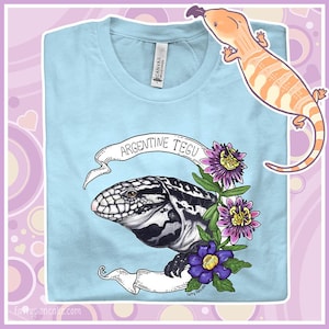 Può includere: Maglietta blu chiaro con un'illustrazione in bianco e nero di una lucertola Tegu argentina con un design floreale. Il testo "Argentine Tegu" è scritto su uno stendardo sopra la lucertola.
