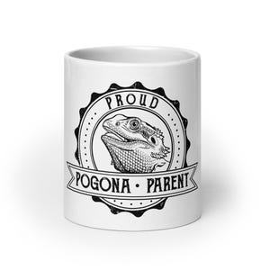 Peut inclure: Un mug en céramique blanche orné d'un motif circulaire noir. Le design comprend une illustration détaillée d'un lézard dragon barbu dans une bordure festonnée, avec le texte PROUD POGONA PARENT imprimé en police noire stylisée.