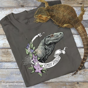 Può includere: Una maglietta grigio scuro con un'illustrazione in bianco e nero di un drago di Komodo con accenti floreali. Il testo "Komodo Dragon" è scritto su uno stendardo bianco.