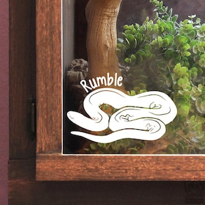 May include: A white vinyl decal applied to a glass surface featuring a stylised snake illustration and the word Rumble written in a curved font above the snake. The decal is positioned on the glass front of a wooden enclosure containing greenery.