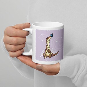 May include: A white ceramic mug featuring a graphic illustration of a tan spotted lizard sitting upright in grass, looking up at a small blue butterfly. The background of the illustration is a soft lavender color with white swirling accents.