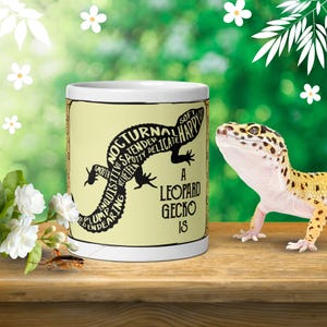 Puede incluir: Una taza de cerámica blanca con una silueta negra de un gecko leopardo rellena de palabras descriptivas como nocturnal, tender y plump, con el texto A LEOPARD GECKO IS impreso en negro junto al gráfico sobre un fondo de color amarillo claro.