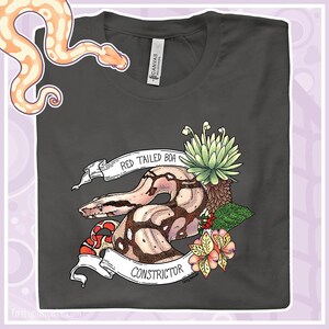 May include: Dark grey t-shirt with a detailed illustration of a Red Tailed Boa Constrictor snake. The design includes a banner with the text "Red Tailed Boa Constrictor", flowers, and foliage. The shirt is made of a soft material.
