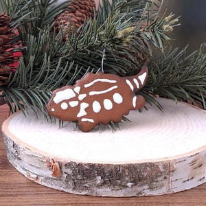 May include: A brown, dinosaur-shaped Christmas ornament with white detailing. The ornament is suspended in front of a pine branch and a wooden slice.