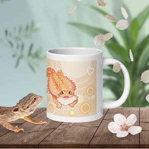May include: A white ceramic mug featuring a cartoon illustration of an orange bearded dragon on a light tan background with circular patterns and small white hearts. The mug has a standard handle and a smooth, glossy finish.