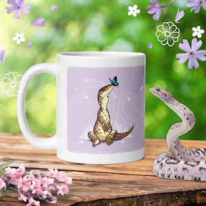 May include: A white ceramic mug featuring a graphic illustration of a brown spotted lizard sitting upright with a small blue butterfly perched on its nose, set against a light purple background with subtle swirling patterns and white grass.