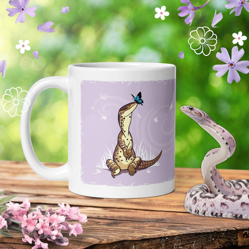 May include: A white ceramic mug featuring a graphic illustration of a brown spotted lizard sitting upright with a small blue butterfly perched on its nose, set against a light purple background with subtle swirling patterns and white grass.