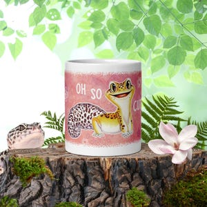 May include: A white ceramic mug featuring a colorful illustration of a yellow and brown spotted leopard gecko on a pink patterned background with the text OH SO CUTE printed in white letters next to the gecko character.