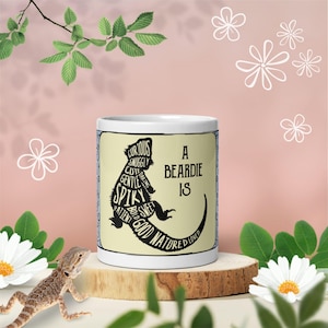 May include: A white ceramic mug featuring a black silhouette of a bearded dragon filled with descriptive words including curious, snuggly, cute, daring, gentle, spiky, sweet, patient, bold, good natured, and loved, with the text A BEARDIE IS.