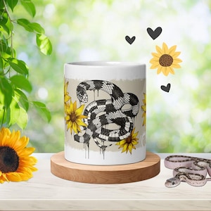 Puede incluir: Una taza de cerámica blanca con una ilustración en blanco y negro de una serpiente enroscada con efectos de tinta goteante, rodeada de girasoles amarillos y pequeños iconos de corazones negros sobre un fondo rectangular beige claro.