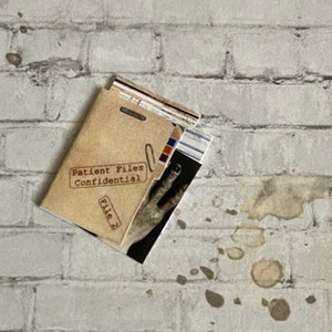 May include: A tan file folder labeled "Patient Files Confidential" with "File 2" printed on it. The folder is slightly open, revealing a black and white image of a hand. The background is a white brick wall with brown stains.