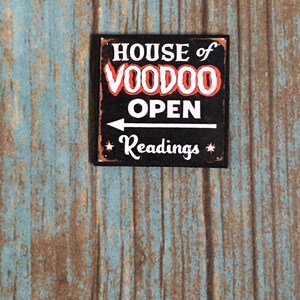 May include: A black sign with white text that reads "HOUSE of VOODOO OPEN" with an arrow pointing to the right and the text "Readings" below the arrow. The sign has a distressed look and is made to look like metal.