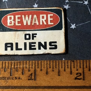 May include: A black and red sign with white text that reads "Beware of Aliens". The sign is made of metal and has a distressed look.