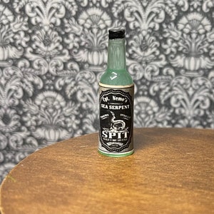 May include: A glass bottle with a green liquid inside. The bottle has a black and white label that reads "Cpt. Nemo's Sea Serpent Infused SPIT". The label also features a black and white illustration of a sea serpent.