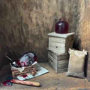 May include: A miniature horror scene featuring a severed head with green eyes and blood, an axe, a jug, and wooden crates. A burlap sack and newspaper complete the unsettling display. The scene is set against a rustic wooden backdrop.