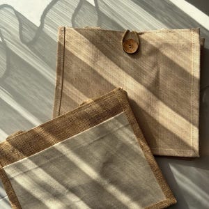 Könnte beinhalten: Zwei beige rechteckige Stofforganizer mit natürlicher Fasertruktur. Einer hat einen Knopfverschluss. Die Organizer liegen auf einer hellen Oberfläche, wobei Schatten von einem Fenster ein Streifenmuster erzeugen.