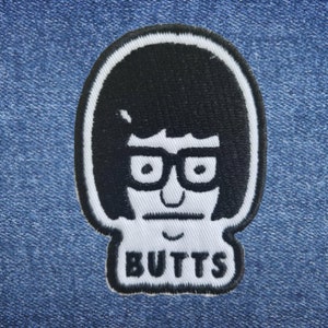 Könnte beinhalten: Schwarz-weiß gestickter Aufnäher mit einer Comic-Frau mit schwarzen Haaren, Brille und einem ernsten Gesichtsausdruck. Der Text "BUTTS" befindet sich unter dem Comic.