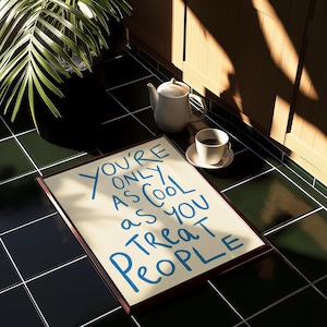 May include: A framed print with the text "You're only as cool as you treat people" in blue lettering on a cream background. The print is sitting on a black and green tiled floor.