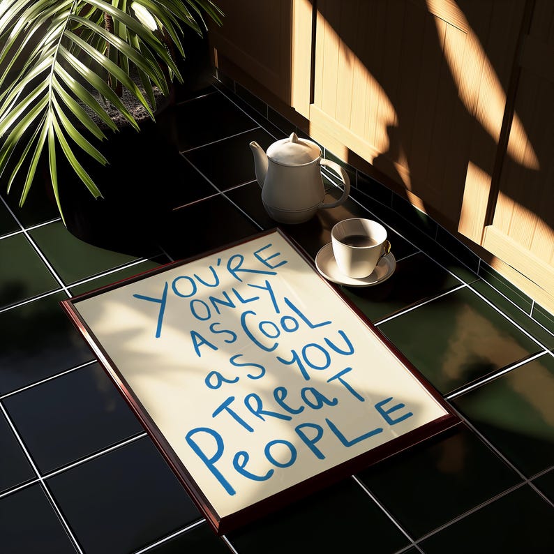 May include: A framed print with the text "You're only as cool as you treat people" in blue lettering on a cream background. The print is sitting on a black and green tiled floor.
