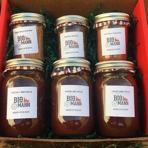 May include: A red gift box filled with six glass jars of Big Mann BBQ sauce and rubs. The jars are labelled with the brand name and the type of sauce or rub. The jars are arranged in two rows of three. The box is sitting on a wooden surface.