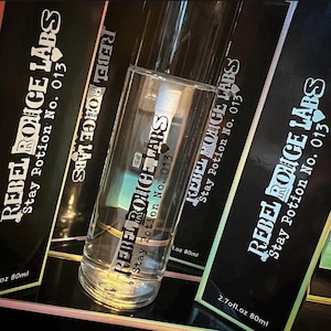 Puede incluir: Una botella de vidrio transparente con una banda dorada, etiquetada como "REBEL ROUGE LABS Stay Potion No. 013." La botella está frente a cajas negras con la misma marca. Las cajas tienen acentos iridiscentes y el texto "2.7 fl.oz 80ml."