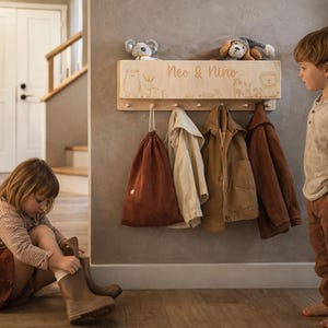 May include: A light-colored wooden wall-mounted coat rack featuring five small pegs and a shelf on top. The front of the shelf is engraved with the text "Neo & Nino" and illustrations of various animals, including a bear, a fox, and a lion.